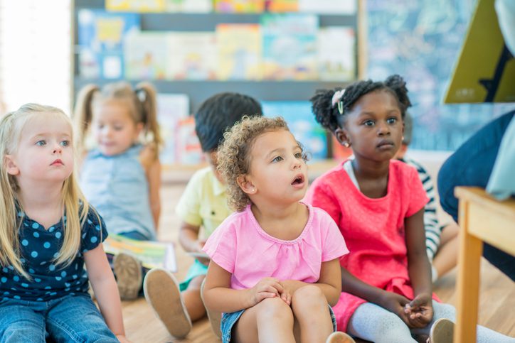 Curly-haired young girl in a pink shirt looking engaged in a classroom setting with other children in the background.