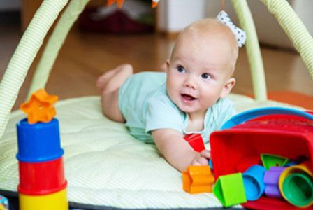Smiling infant lies on a soft white pillow, surrounded by colorful stacking cups for sensory and motor skill development.