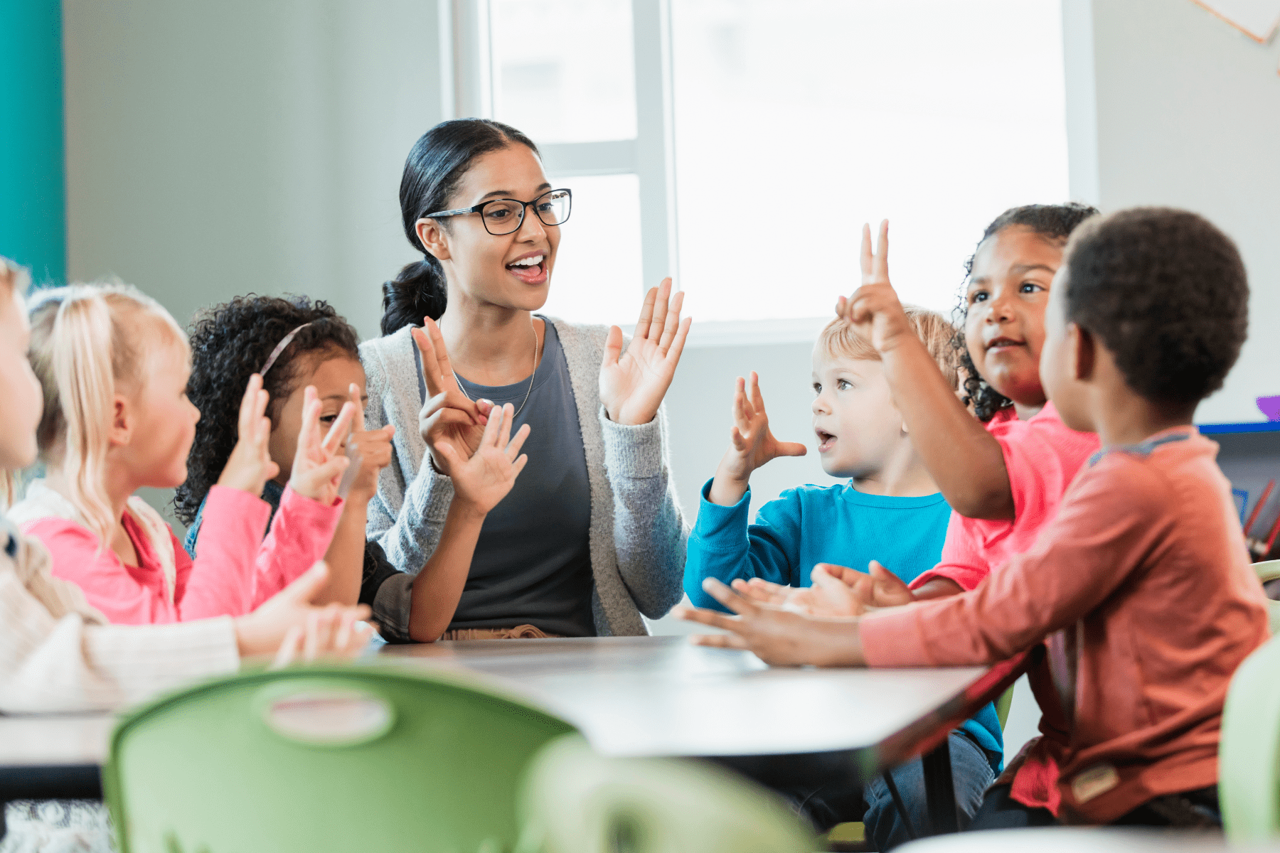 A smiling teacher engages with young children in a fun classroom activity, using hand gestures as they participate together.