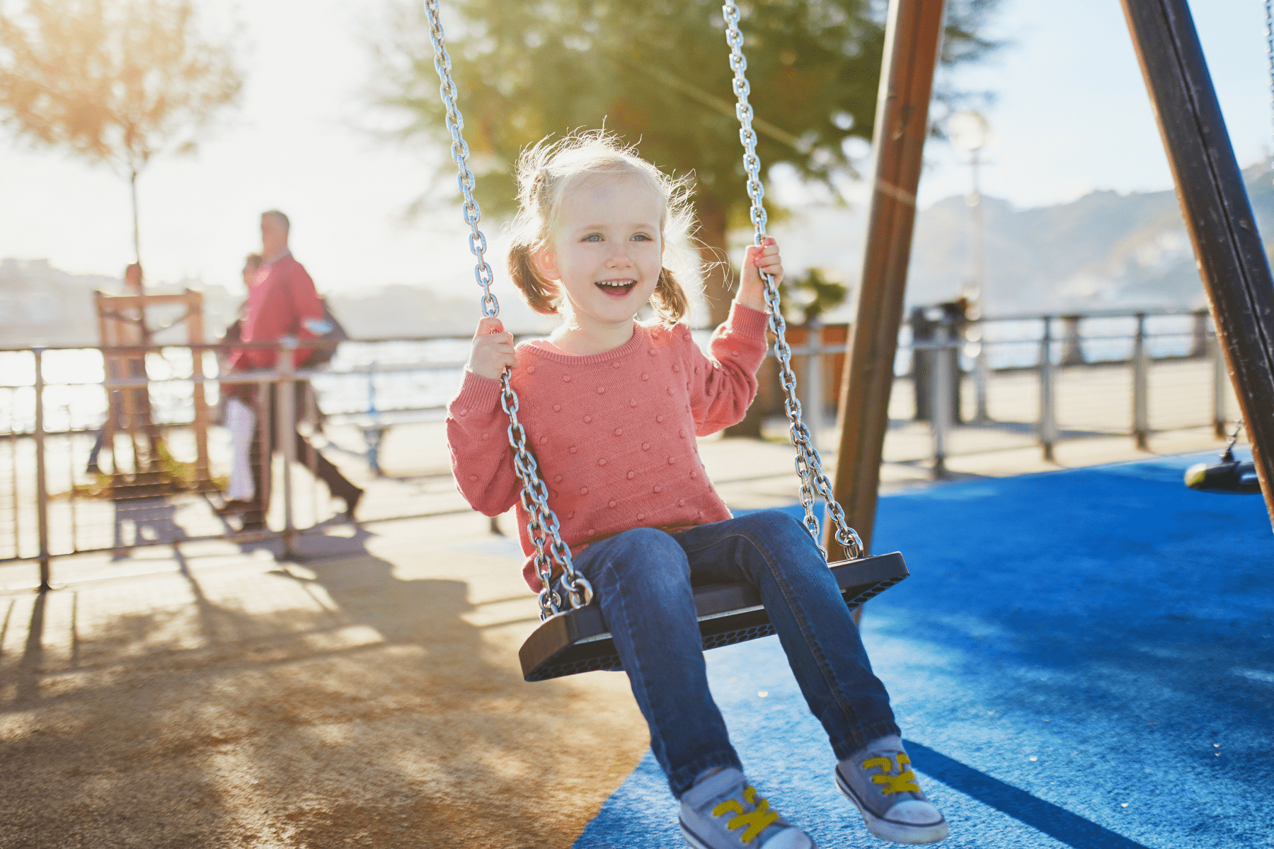Smiling young girl with pigtails in a pink sweater swinging at a sunny outdoor playground with blue flooring.