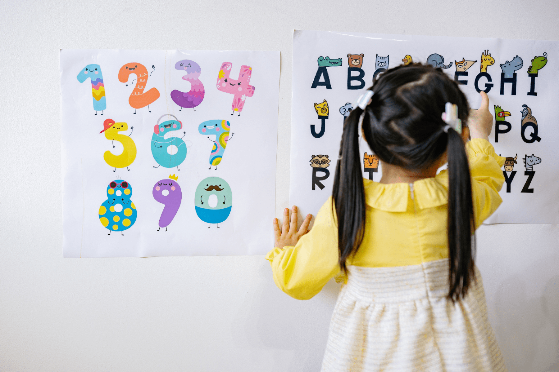 A young girl pointing at an alphabet poster, next to a colorful number poster