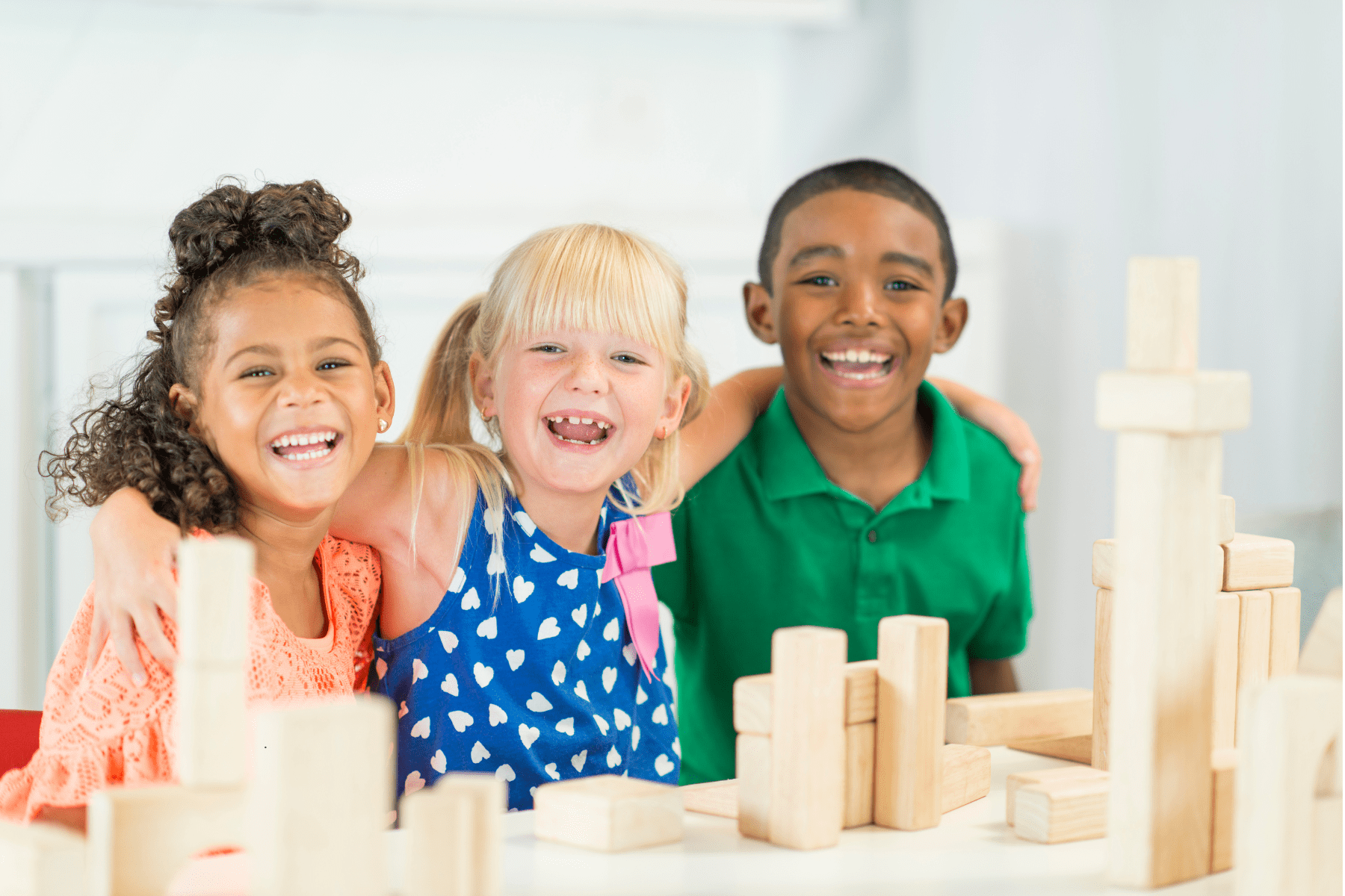 Three smiling children with arms around each other sit at a table building wooden block towers in a bright classroom.
