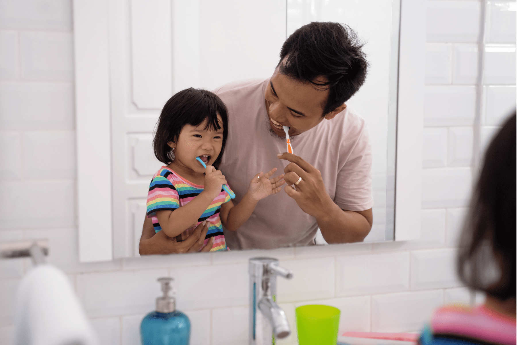 Father and young daughter smiling while brushing their teeth together at the bathroom sink in front of a mirror.