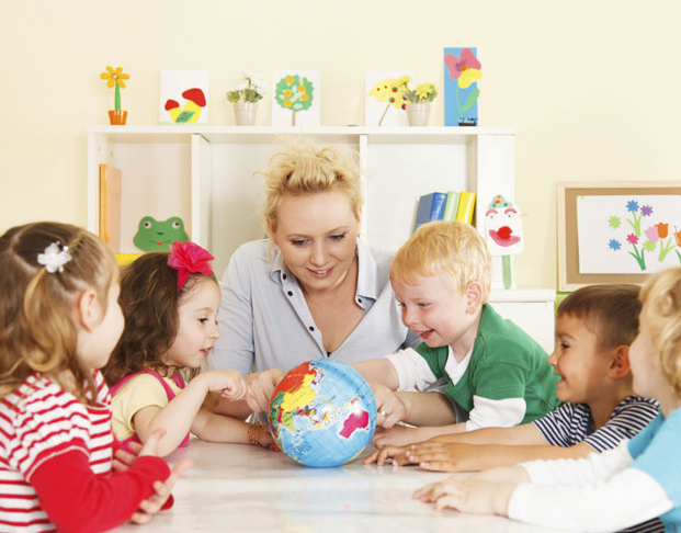 Daycare class sitting at a table with the teacher all looking at a Globe.
