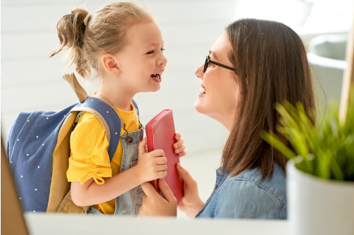 3-year-old girl in a yellow shirt with a backpack on handing her book to Daycare Teacher.