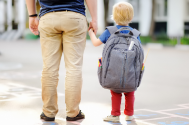A preschooler with a big grey backpack holding his Father’s hand.